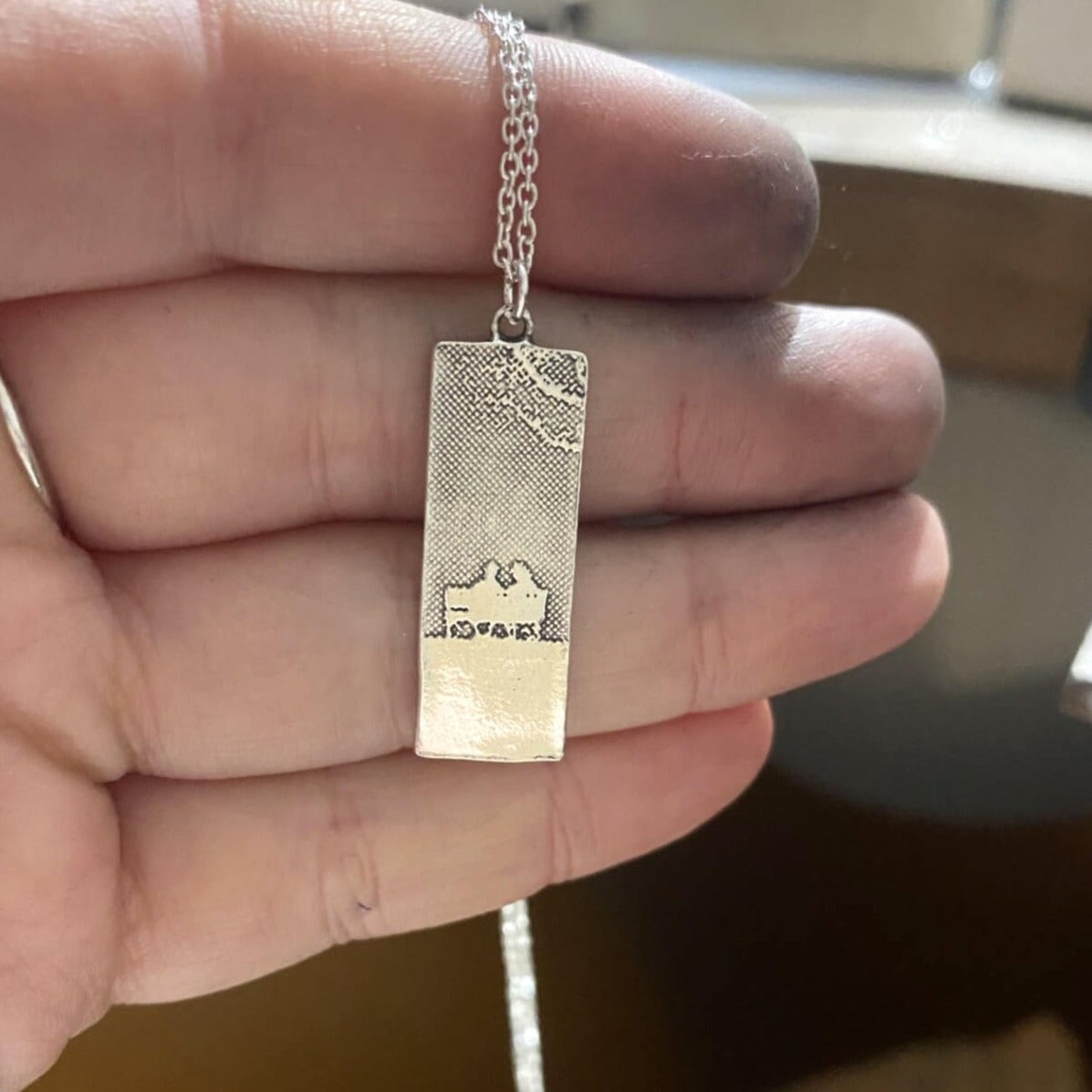 Sterling silver always with you memory necklace held in makers hand with etched details of two figures on a bench.