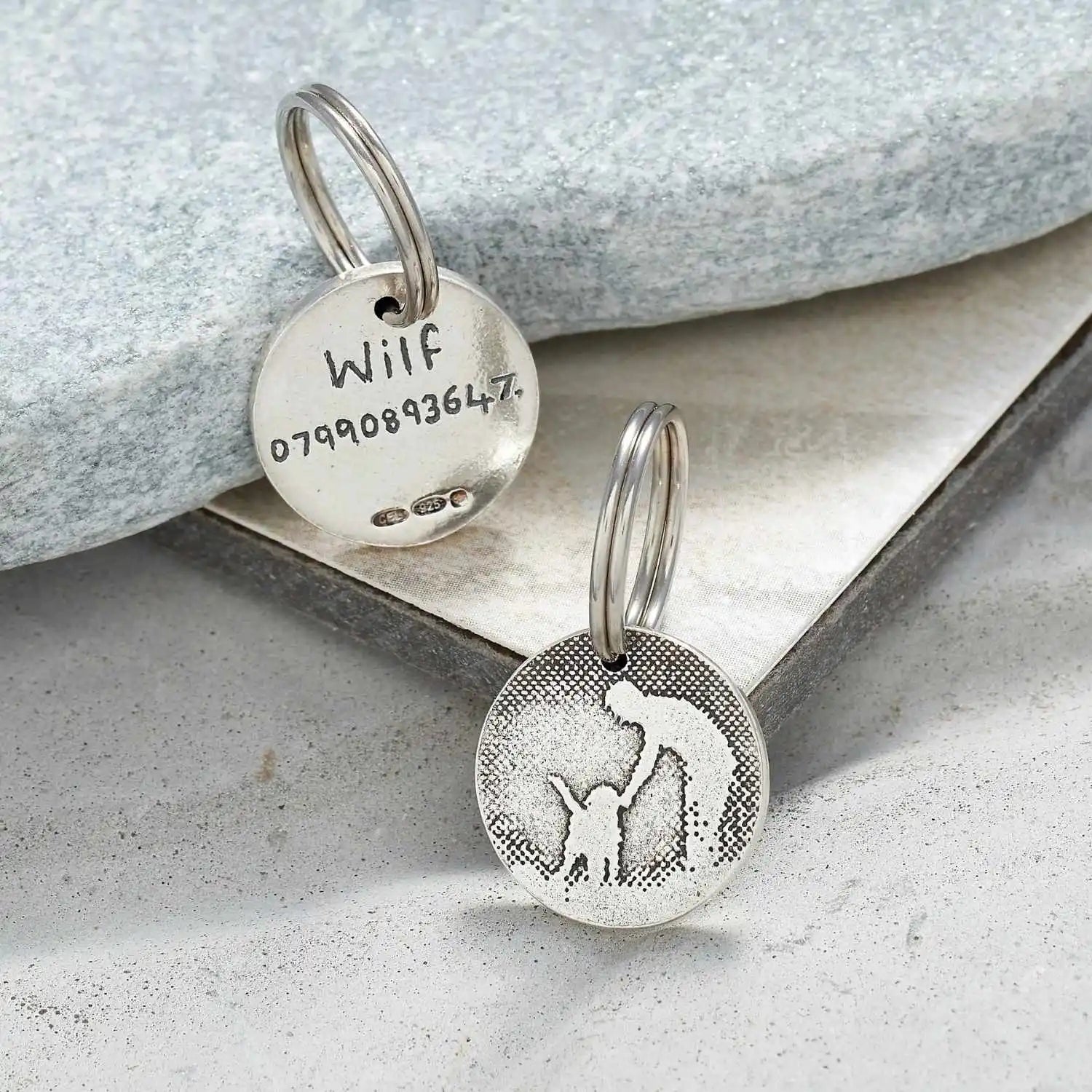Two round sterling silver pet ID tags with split rings, one engraved with a name and phone number and the other featuring a silhouette of an adult and child, displayed on a light gray stone surface.