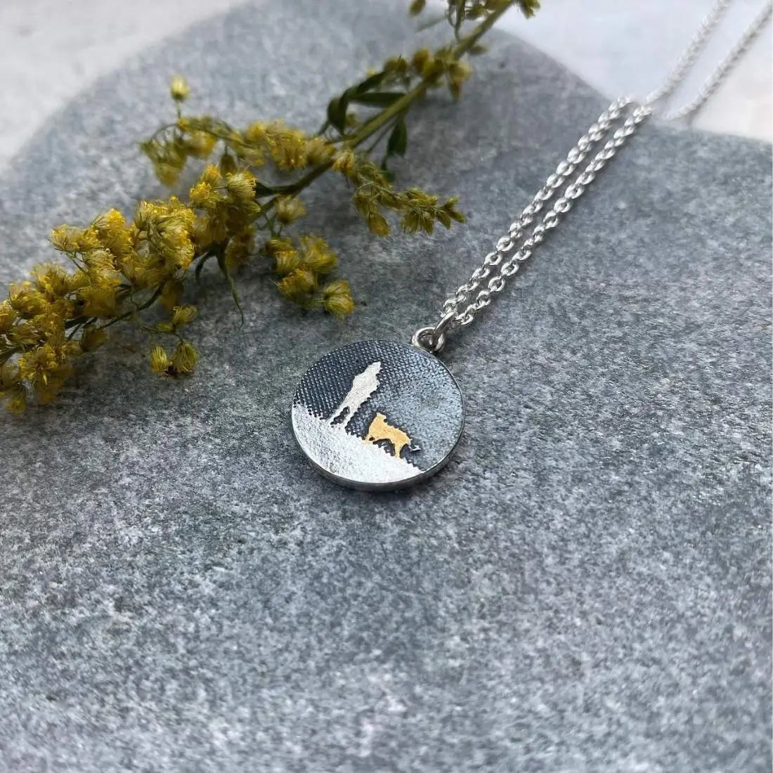A dog necklace featuring a dog and it's owner beneath an oxidised silver sky. The person and the ground are silver and the dog is finished with 22ct gold vermeil.