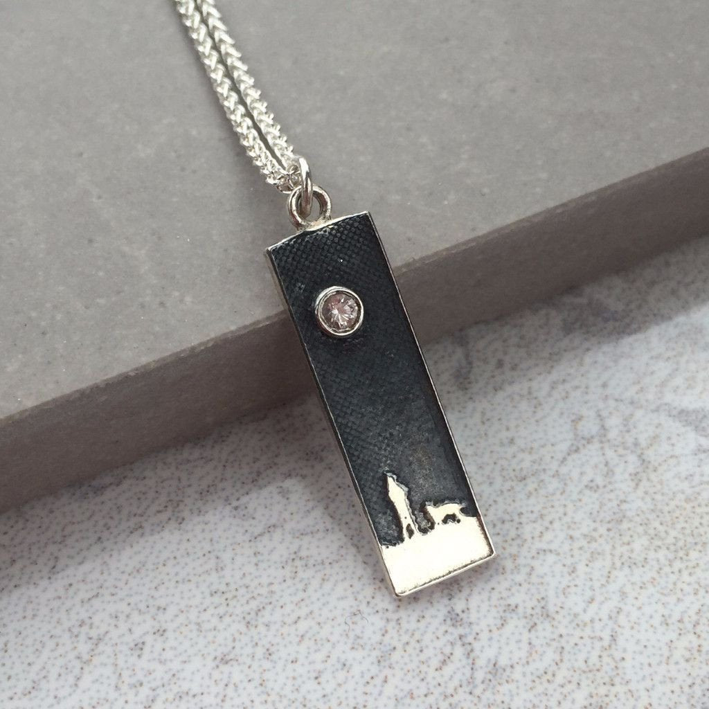 Side by Side Beneath the White Sapphire Sky necklace displayed on a grey tile background showing the etched figures and white sapphire moon detail.