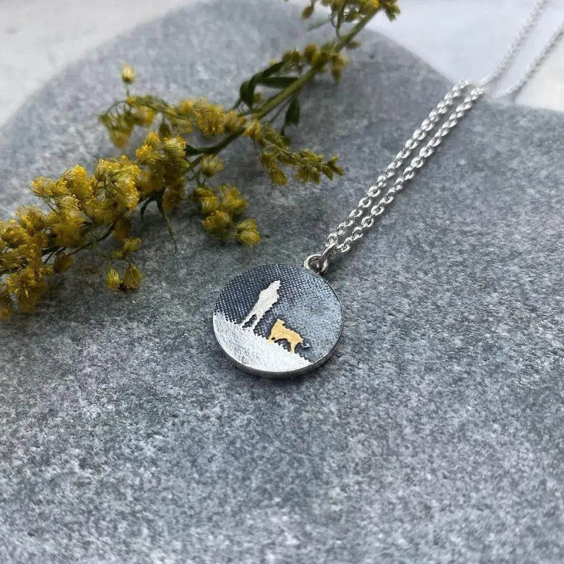 Walks Under the Night’s Sky gold dog necklace on a textured grey background with green foliage.