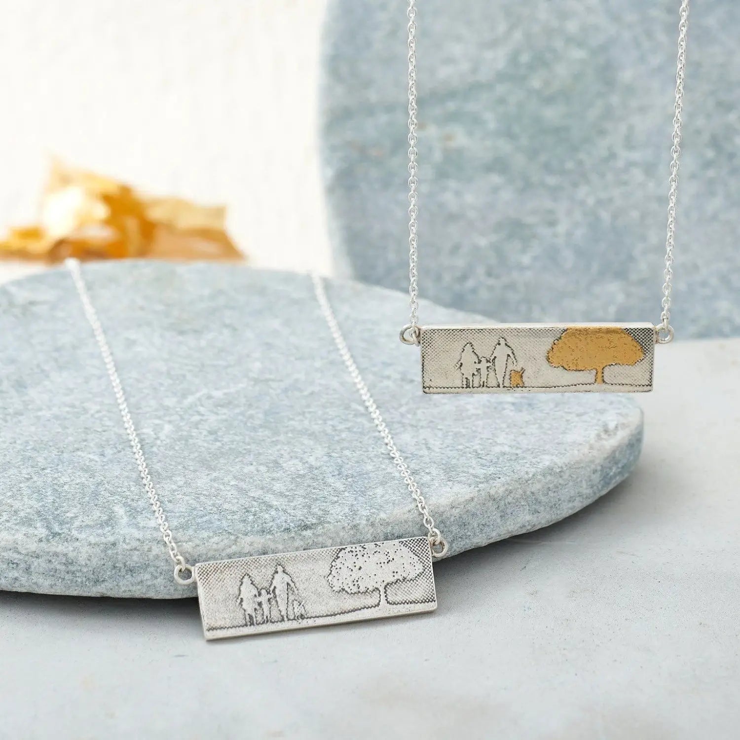 Lifestyle image showing the sterling silver and gold‑detail Dog Lovers Family Tree Necklaces, each featuring an etched family of three with their dog beneath a detailed tree in a rectangular landscape design.