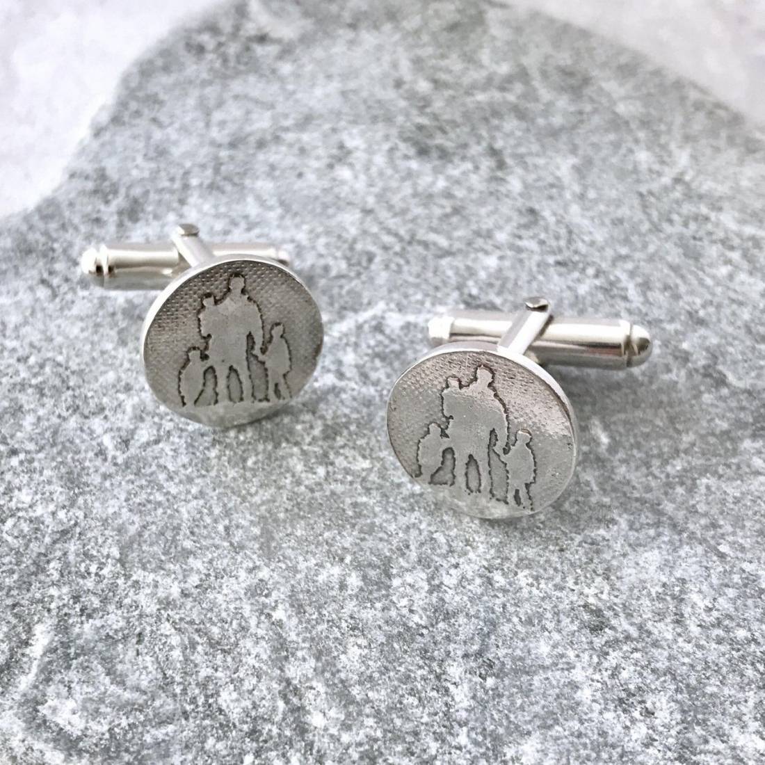 A pair round silver cufflinks with engraved silhouettes of a father and his three children, displayed on a textured gray stone surface.