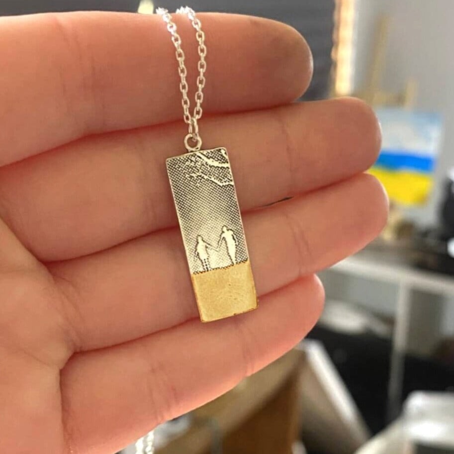 Hand in Hand Beach Memories Necklace in makers hand showing two figures walking hand in hand along the shoreline, etched into sterling silver.