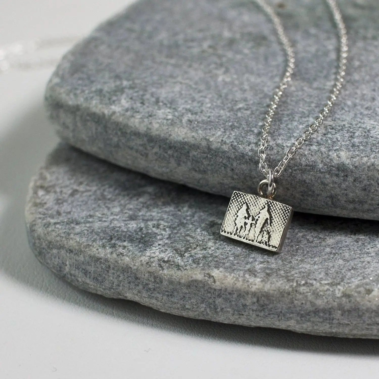 Close‑up lifestyle image of the Little Dog Lovers Family Necklace showing the etched family of three and their dog in a petite sterling silver rectangular pendant with oxidised detail.