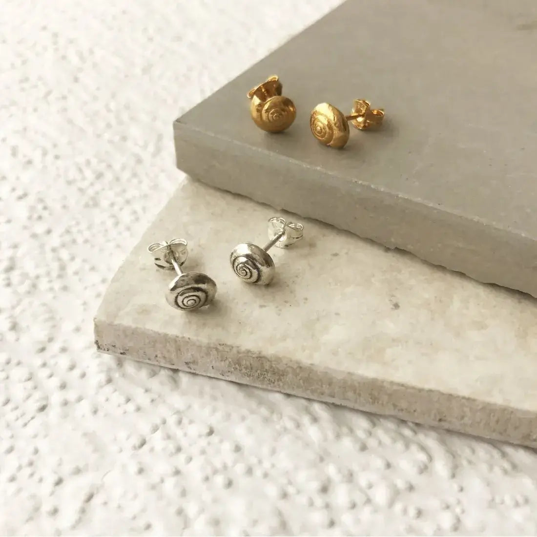 Flat-lay image showing the little round gold sea shell earrings alongside the polished sterling silver version on stepped tiles.