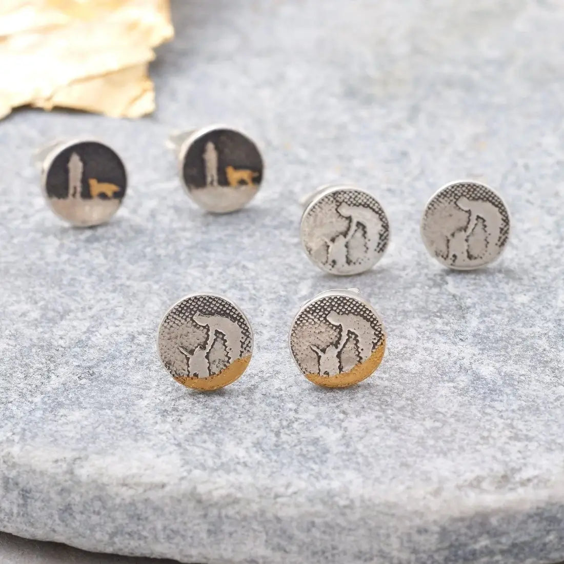 Lifestyle image showing three pairs of Night’s Sky Dog Stud Earrings: the gold‑detail version in the background, the silver petting‑scene version in the middle, and the beach‑scene petting version in the foreground.