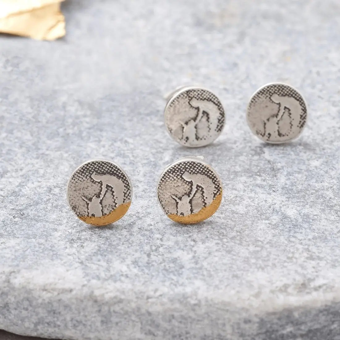 Lifestyle image showing round dog stud earrings, with the beach‑scene version in the foreground and the silver version behind, each depicting an owner leaning down to pet their dog.