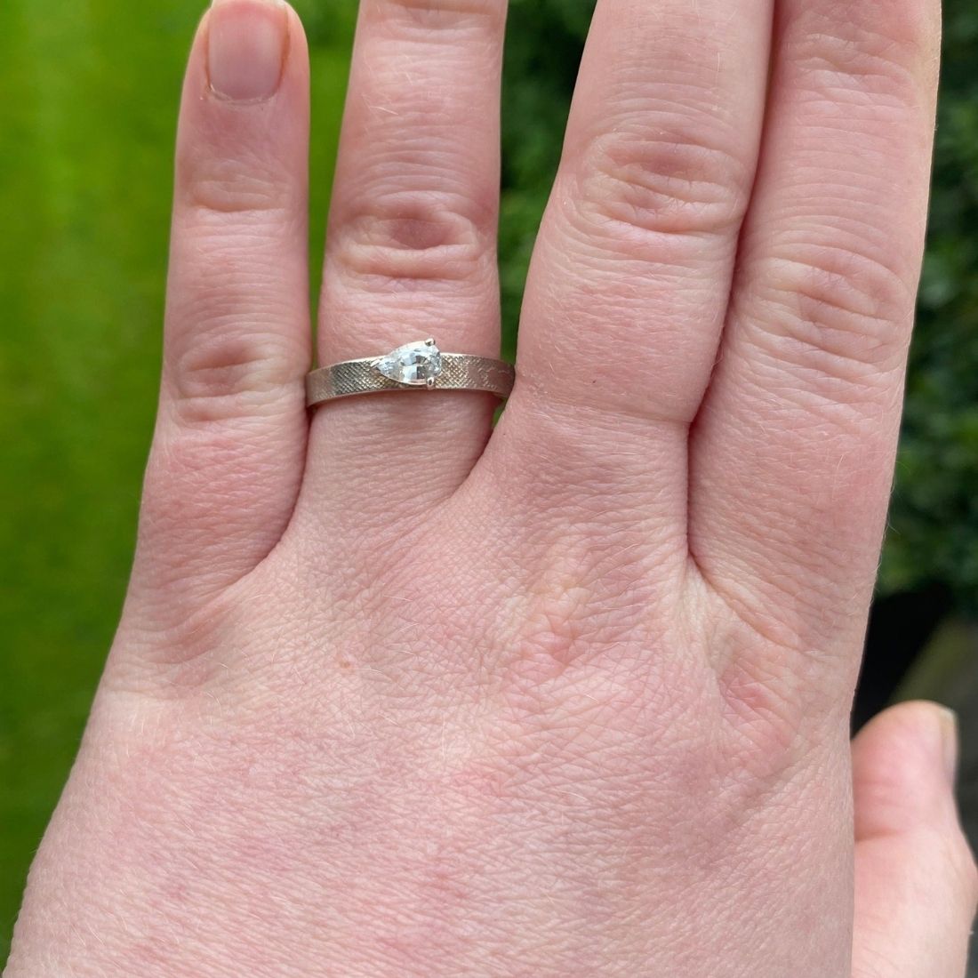 White gold or platinum engagement ring with textured band and pear‑shaped diamond on models hand.