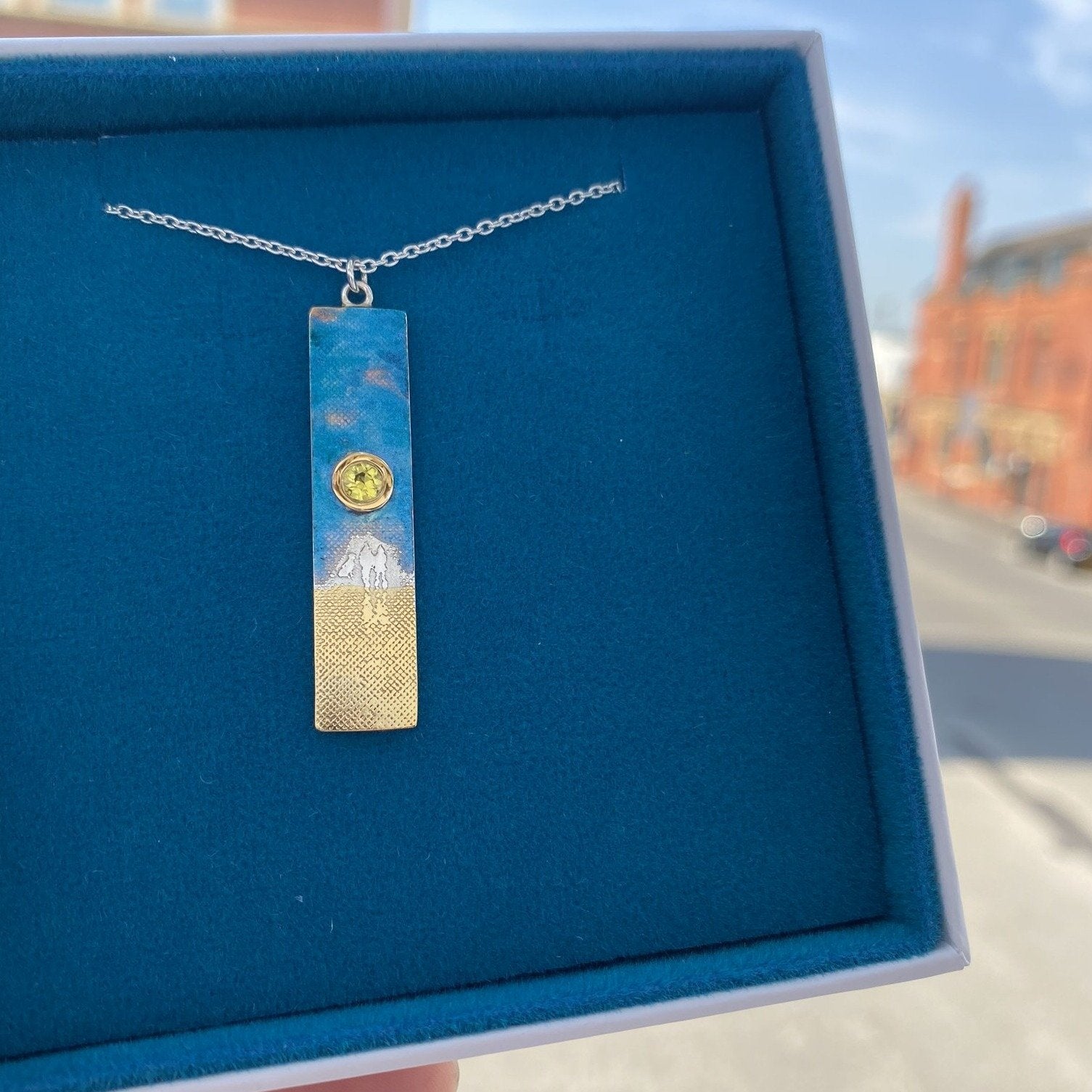 Sterling silver sunset couple and dog necklace featuring etched figures walking with their dog, a blue enamel sky, a yellow sapphire sun and 22ct gold vermeil sand detail.