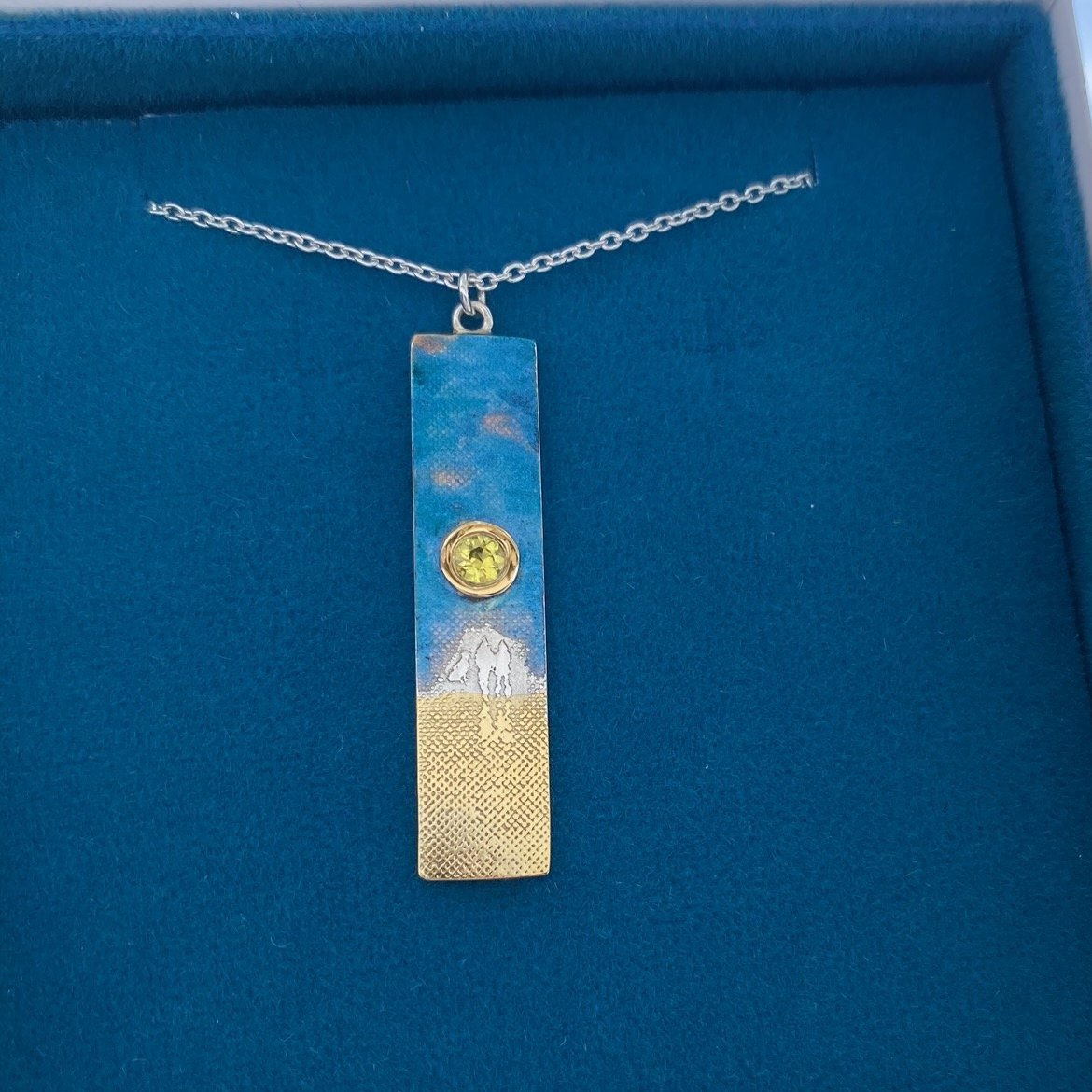 Close-up of the sunset couple and dog necklace showing the etched figures, blue enamel sky, yellow sapphire sun and 22ct gold vermeil sand detail.